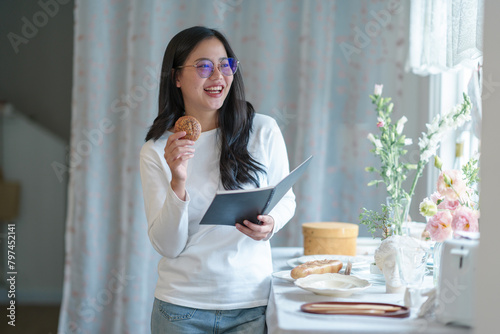 Attractive young woman wearing glasses holding a reading book and snacks stands smiling relaxedly in a bakery, shop, coffee shop in the morning. lifestyle concept working holiday. photo