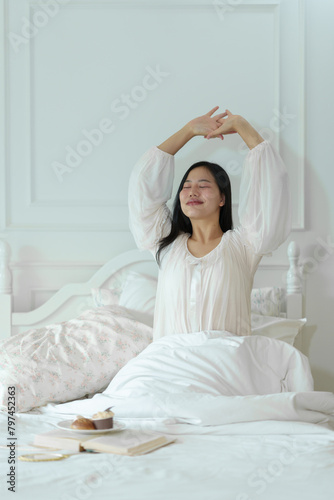 Beautiful Asian woman in white pajamas stretching after waking up in the morning on a comfortable white bed at home in the morning. Holiday lifestyle concept.