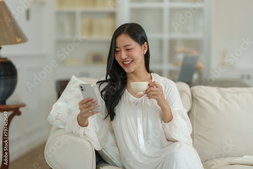 Asian woman in white pajamas sitting on the sofa looking at mobile phone online, holding a cup of hot coffee. Smiling happily in her living room on a relaxing morning. Holiday concept, lifestyle.