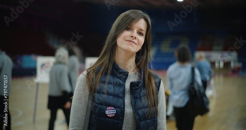 Happy female US citizen after voting during Presidential Election in the United States. Portrait of Caucasian woman with badge smiling and looking at camera standing at polling station. Civic duty.