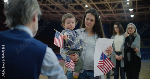 Woman with baby on hands talks with polling officer and takes bulletin. Female American citizen comes to vote in polling station. Political races of US presidential candidates. National Election Day.