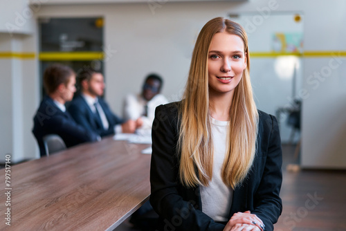 Confident blonde businesswoman in smart casual attire leads meeting, sits at conference table with diverse team in background. Professional office setting, teamwork, leadership, modern workplace. photo