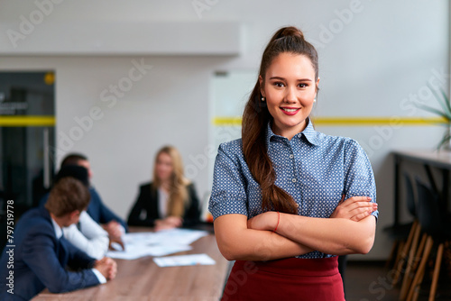 Confident asian exec woman stands in modern office, arms crossed, smiling at camera, team in discussion behind her, embodying female leadership, pro success in a smart casual attire. photo