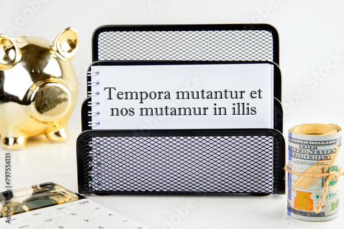 Tempora mutantur et nos mutamur in illis Translated from Latin, it means Times are changing, and we are changing with them. on a white notepad with money and a piggy bank photo