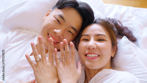 Top view young couple two asia people lying down on bed fun video phone call relax smile look at webcam camera talk show ring on hand finger. Sweet lover asian man woman on family day love life begin.