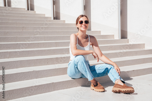 Beautiful smiling model in sunglasses. Female dressed in summer hipster white T-shirt and jeans. Posing near white wall in the street. Funny and positive woman having fun outdoors, sits at stairs