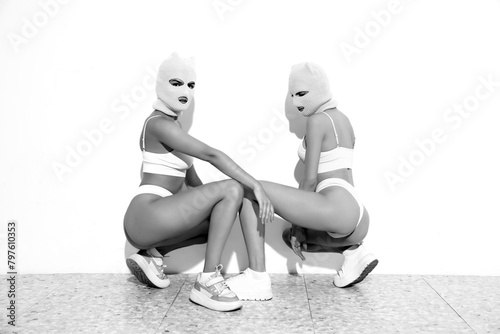 Two beautiful sexy women in underwear. Models wearing bandit balaclava mask. Hot seductive female in nice lingerie posing near white wall in studio. Crime and violence. Sits on floor. Black and white