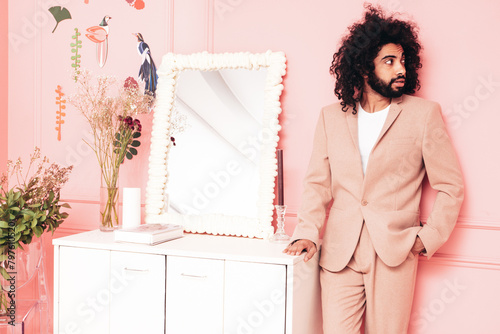 Handsome smiling hipster  model. Unshaven Arabian man dressed in elegant beige suit . Fashion male with long curly hairstyle posing near pink wall in pure interior with flowers in studio