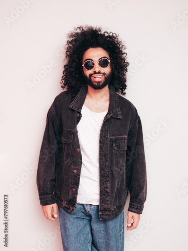 Handsome smiling hipster  model. Sexy unshaven Arabian man dressed in summer stylish black jacket and blue jeans. Fashion male with long curly hairstyle posing near grey wall in studio