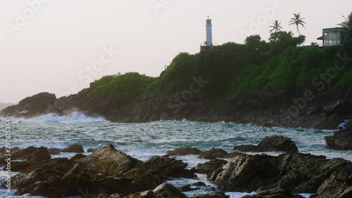 Lighthouse stands on green hill above ocean waves crashing against rocky shore at sunset. Tranquil dynamic nature, meditation, scenic backgrounds. Sunlight glistens water, serene dusk coast landscape. photo