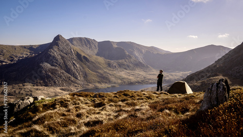 An explorer and photographer on a mountain summit overlooking a spectacular valley from his wild camping tent