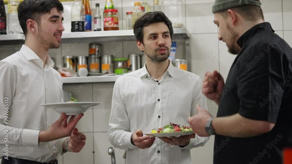 Confident and joyful waiters communicate with the chef about the salads he prepared in the kitchen in a restaurant. Guys in white shirts talk about salads with the chef who prepared them in the Confident and joyful waiters communicate with the chef about the salads he prepared in the kitchen in a restaurant. Guys in white shirts talk about salads with the chef who prepared them in the