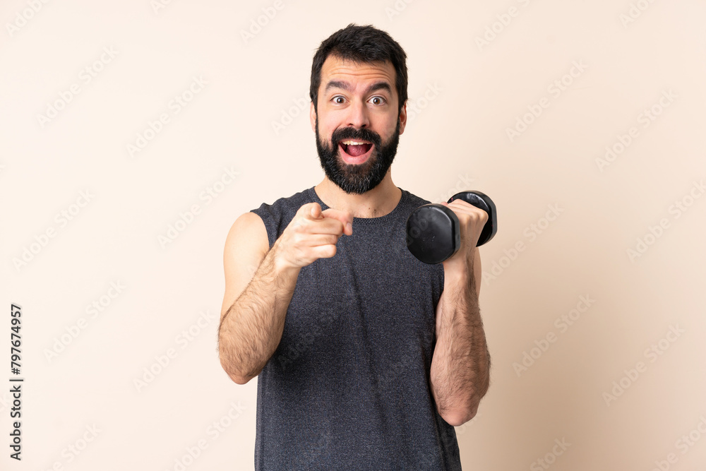Caucasian sport man with beard making weightlifting over isolated background surprised and pointing front Caucasian sport man with beard making weightlifting over isolated background surprised and pointing front