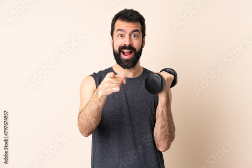Caucasian sport man with beard making weightlifting over isolated background surprised and pointing front © luismolinero