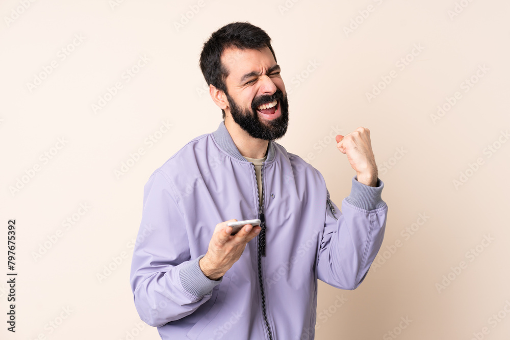 Caucasian man with beard wearing a jacket over isolated background with phone in victory position Caucasian man with beard wearing a jacket over isolated background with phone in victory position