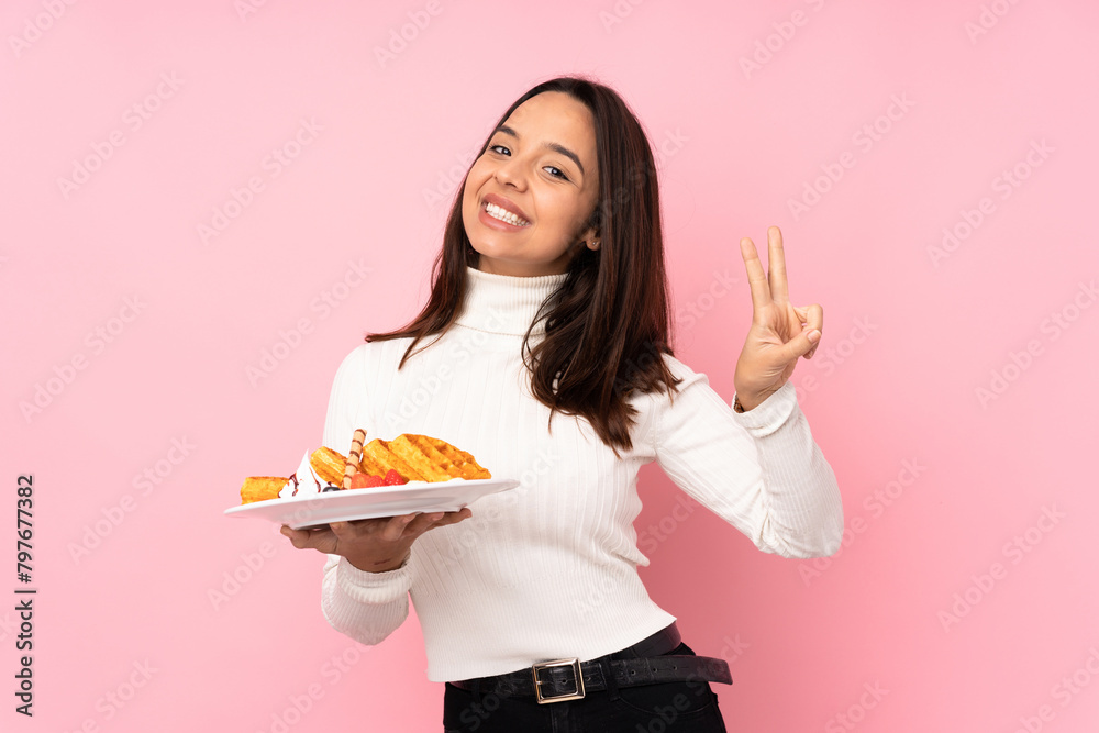 Young brunette woman holding waffles over isolated pink background showing victory sign with both hands Young brunette woman holding waffles over isolated pink background showing victory sign with both hands