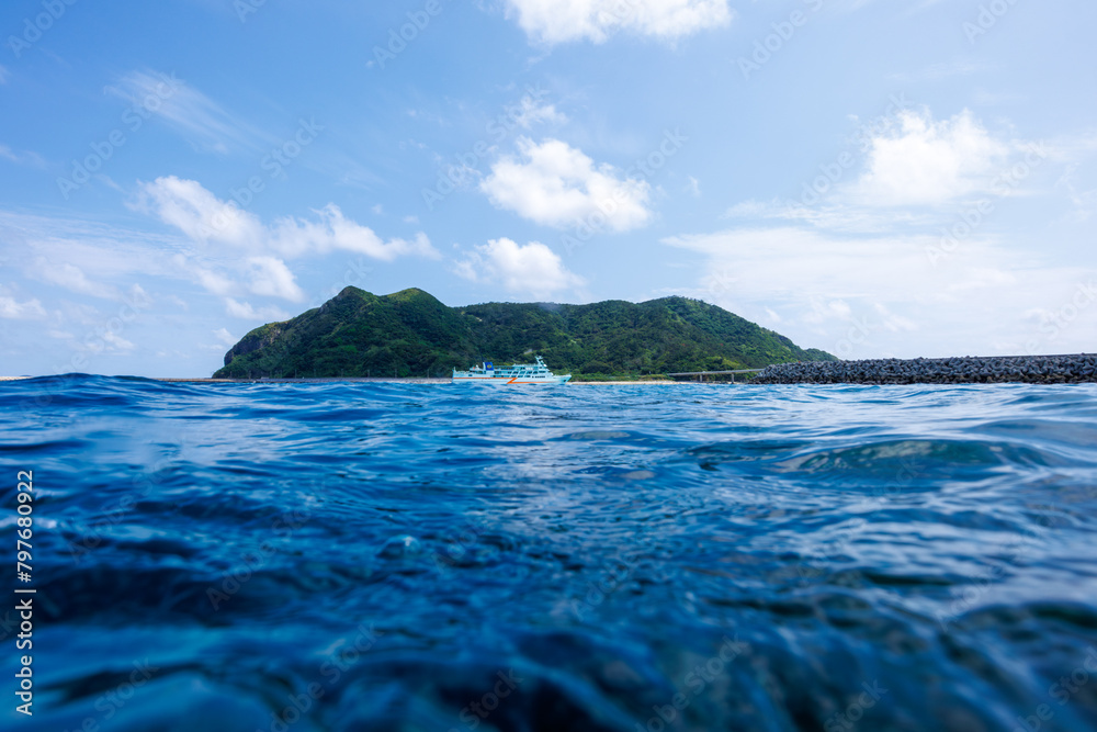 Obraz premium 素晴らしいサンゴ礁の水面から見る、慶良間諸島フェリーのクイーンざまみ3世号と慶留間島の美しい半水面撮影。 圧倒的に大規模な素晴らしく美しいサンゴ礁。沖縄県島尻郡座間味村阿嘉島の阿嘉ビーチにて。 2021年4月28日水中撮影。 A beautiful half-water shot of the Kerama Islands ferry Queen Zamami III and Geruma I