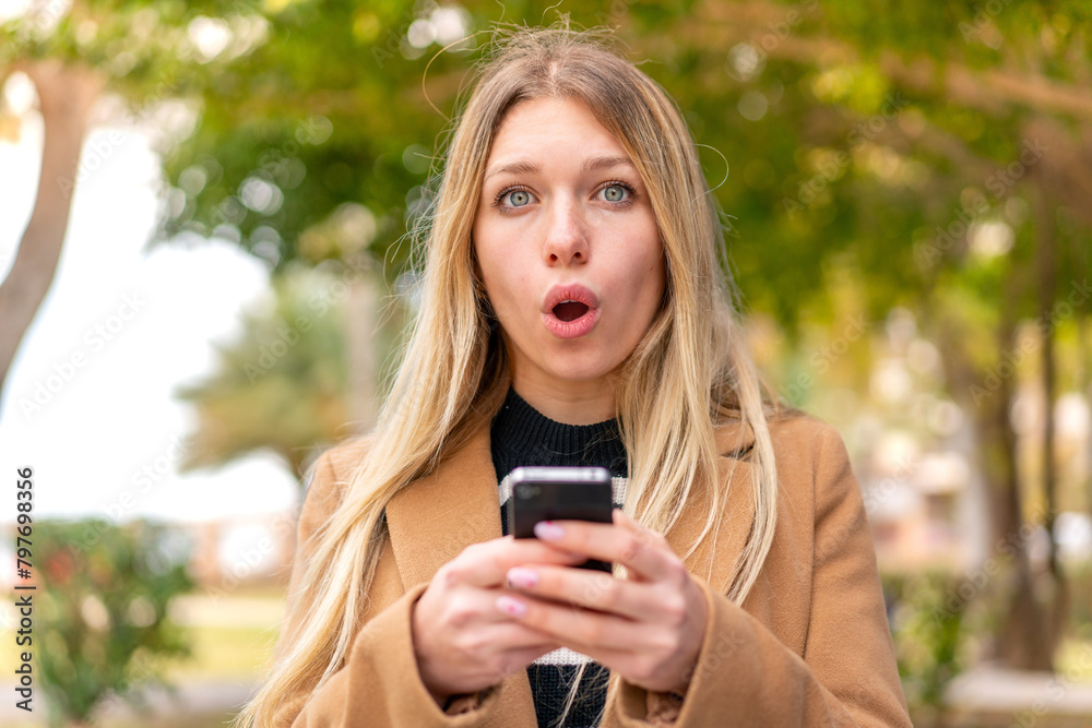 Young pretty blonde woman at outdoors looking at the camera while using the mobile with surprised expression
