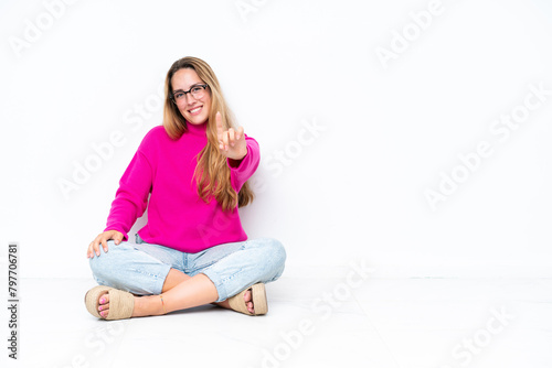 Young caucasian woman sitting on the floor isolated on white background showing and lifting a finger