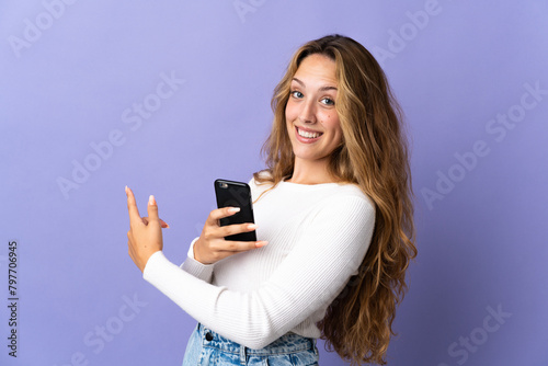 Young blonde woman isolated on purple background using mobile phone and pointing back