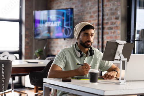 Asian young professional man wearing headphones, holding a pen, looking at a tablet in a modern busi photo