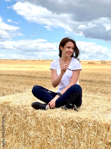Happy young woman model sitting on a haystack, harvest time. Countryside girl on high haystack on the field.