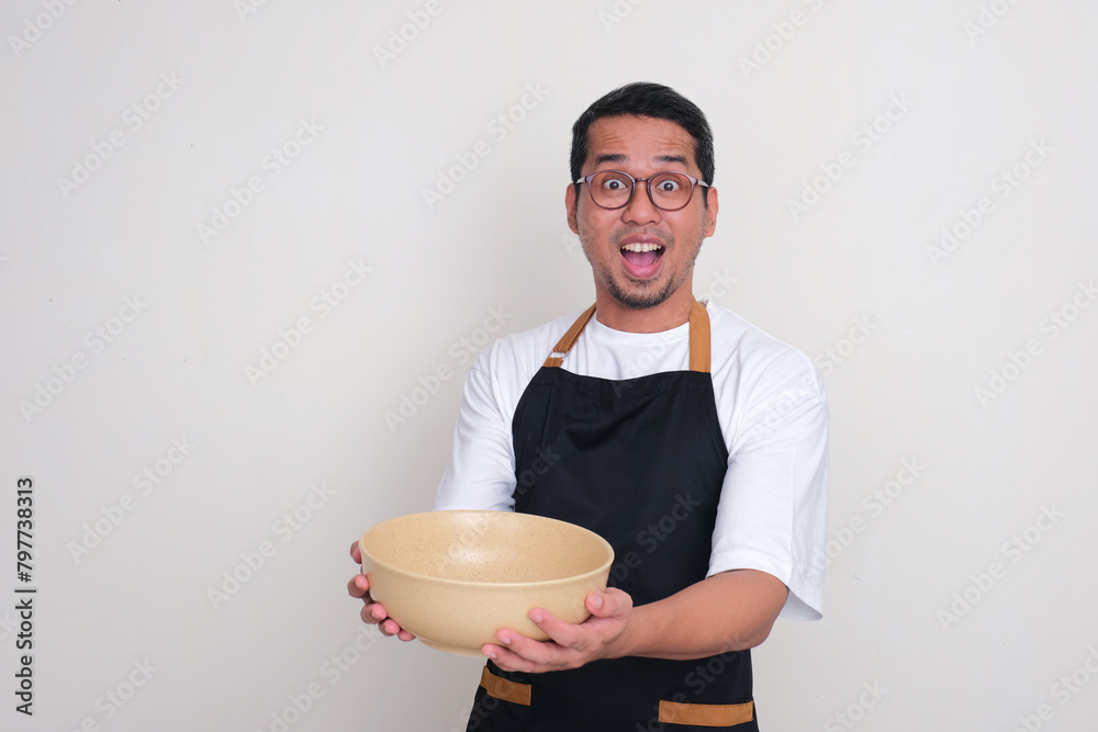 An Asian man wearing apron showing excited expression holding empty big serving bowl
