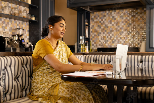Indian woman in yellow sari, smiling while typing on laptop on video call photo
