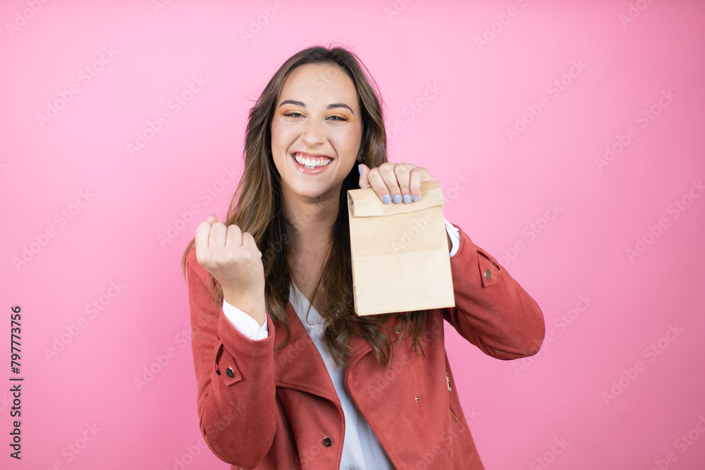 Young beautiful woman wearing casual jacket over isolated pink background holding a paper bag very happy and excited making winner gesture with raised arms, smiling and screaming for success Young beautiful woman wearing casual jacket over isolated pink background holding a paper bag very happy and excited making winner gesture with raised arms, smiling and screaming for success