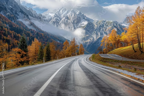 The road curves through a picturesque autumn scene with golden trees and imposing snowy mountains in the backdrop
