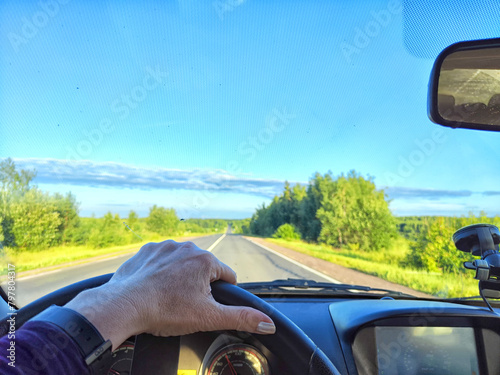Car salon, windshield, hand of woman on steering wheel and landscape. View from seat of driver on nature with Road, trees, blue sky at sunny day. Single trip of female traveler. Partial focus