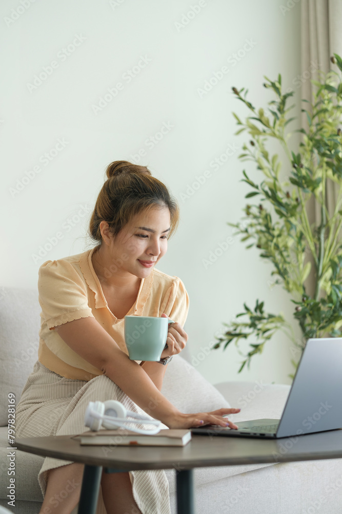 Attractive young Asian woman drinking hot coffee and using laptop in cozy living room. Attractive young Asian woman drinking hot coffee and using laptop in cozy living room.
