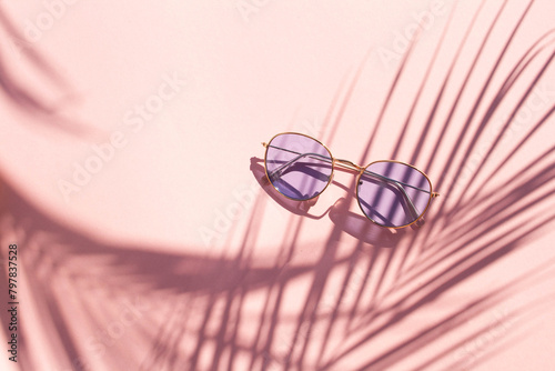 Blue glasses on a pink background with contrasting shadows of a palm tree. Summer vacation concept