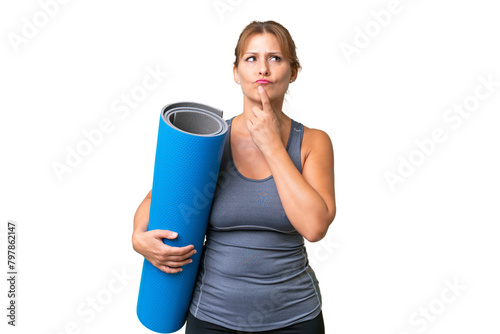 Middle-aged sport woman going to yoga classes while holding a mat over isolated background having doubts while looking up