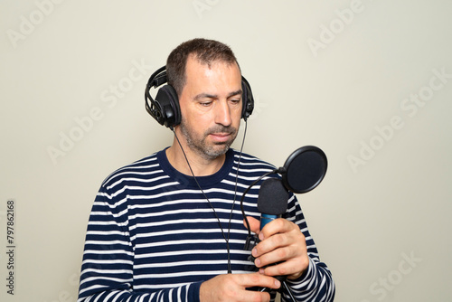 Bearded Hispanic man in his 40s wearing a striped sweater looking confused while inspecting a microphone and headphones, isolated on beige background.