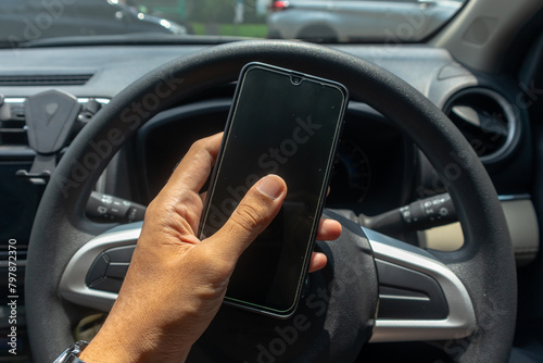 Male hand holding a smartphone with a car steering wheel in the background. Driving while using a smart phone concept. © fadfebrian
