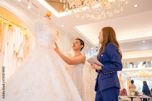 Asian bride is smiling while choosing wedding dress in modern wedding salon, Attractive designer girl using tape meter fitting on wedding dress at wedding studio.