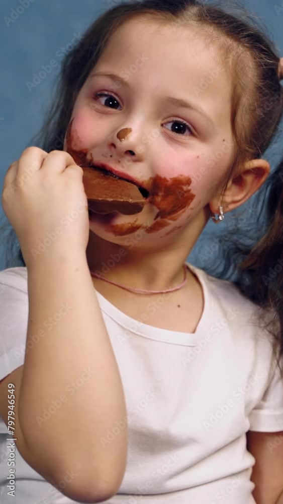 Little girl with dirty face eats bar of chocolate. Funny child with two ponytails is biting tasty black chocolate on blue studio background. Vertical video Little girl with dirty face eats bar of chocolate. Funny child with two ponytails is biting tasty black chocolate on blue studio background. Vertical video
