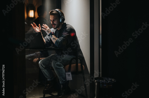 Man with headphones gesturing while gaming at night, displaying emotion and focus, in a moody lit room.