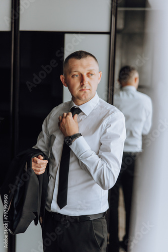 A man in a suit and tie is standing in front of a mirror, adjusting his tie