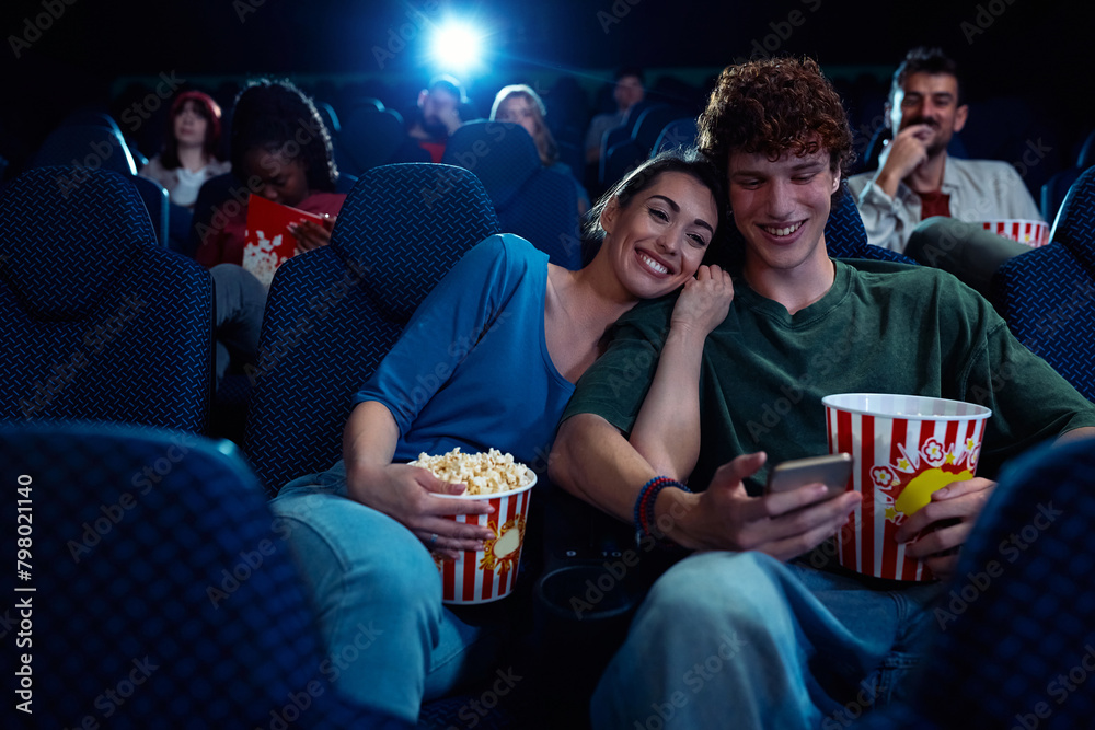 Happy couple using cell phone during movie projection in cinema.