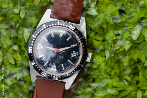 A sturdy outdoor watch, used and waterproof, lies on green plants in the creek, washed by the clear water of nature.