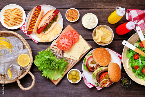 Summer BBQ or picnic table scene with hamburgers, hotdogs, salad and snacks. Above view over a dark wood background.