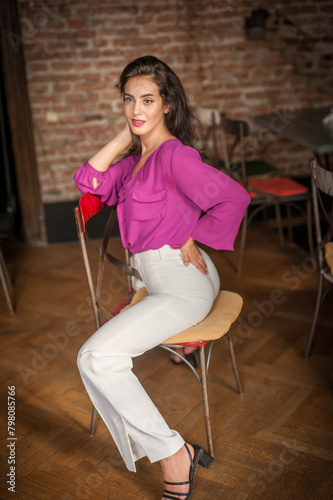 Obraz na płótnie Beautiful young brunette woman in a purple blouse and white pants posing indoor against a brick wall. Portrait of a beautiful young brunette woman w salonie