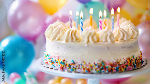 Birthday cake with lit candles and colorful sprinkles  with blurred balloons in the background. Celebration and party concept with copy space.