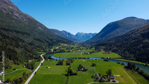 An aerial view of the Norwegian town Stryn, nestled in a lush valley with the iconic, winding Stryn River, flanked by verdant fields and framed by majestic mountains under a clear blue sky. photo