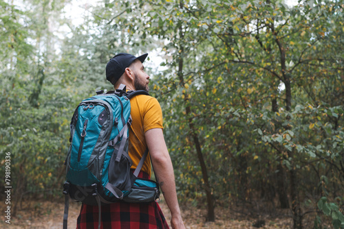 Young man with tourist backpack walking alone in early autumn forest and looking to the side