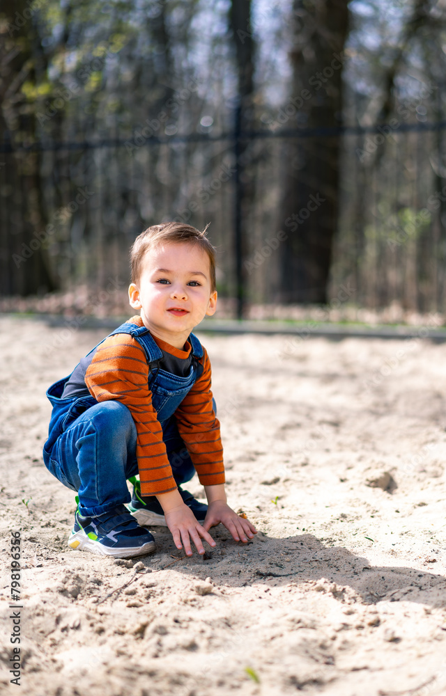 A young boy is crouching in the sand, wearing a blue overalls and a striped shirt A young boy is crouching in the sand, wearing a blue overalls and a striped shirt