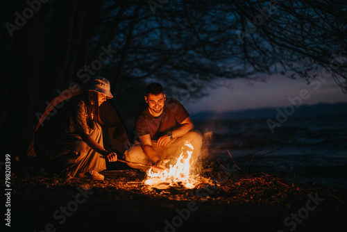 Two friends share a joyful moment outdoors while camping by a lake, cooking dinner over a campfire at night.
