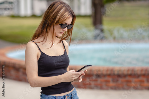 Young woman looking at cell phone in the park with a fountain behind her, horizontal, close up